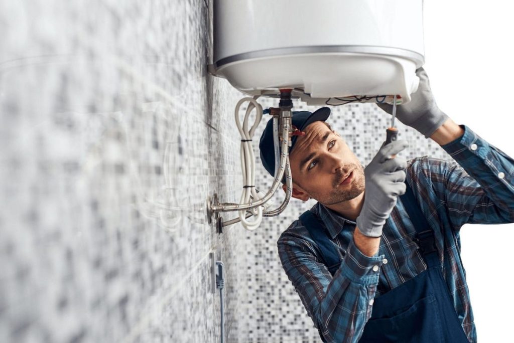 Male plumber wearing gloves, a plaid shirt, and overalls, using a screwdriver to service or install a wall-mounted water heater in a tiled bathroom