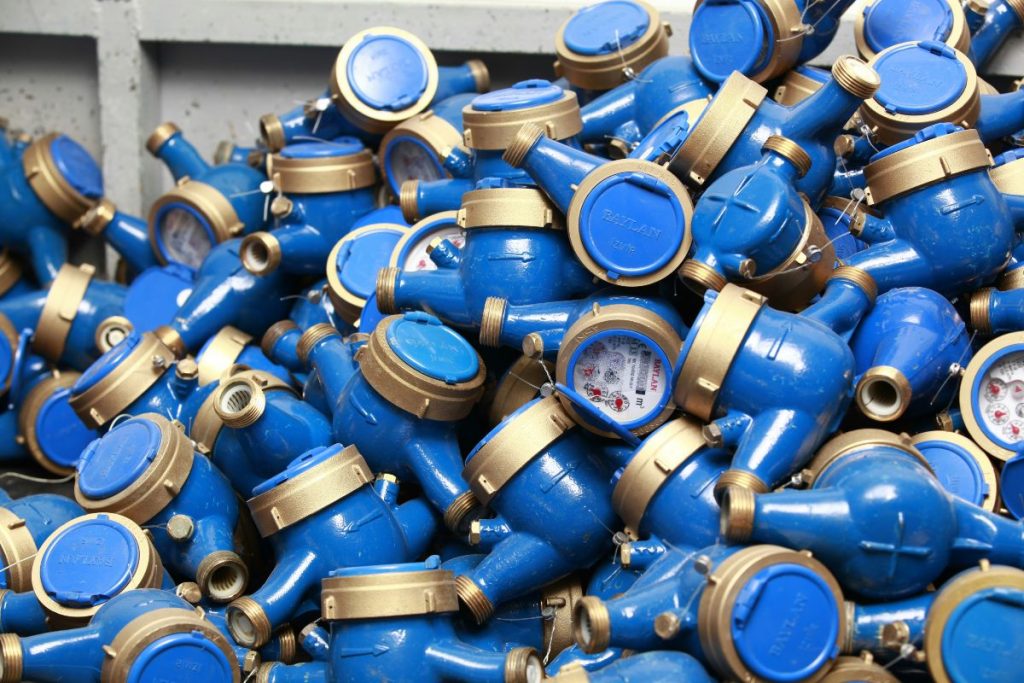 Pile of blue and brass residential water meters, some showing measurement dials and others with protective caps